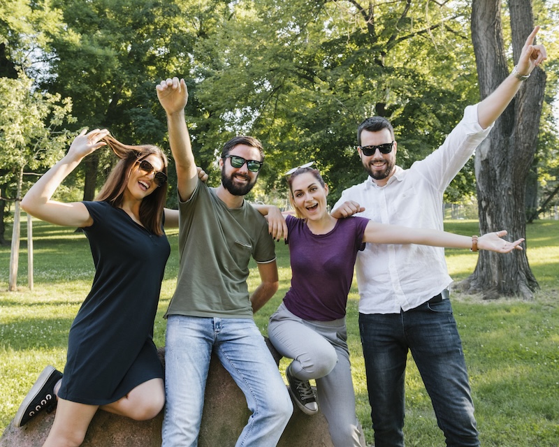 Gruppe fröhlicher Freunde im Freien, die zusammen Spaß haben, in einem sonnigen Park mit grünen Bäumen.